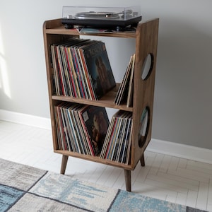 Op de afbeelding: Een houten platenspelerstandaard in mid-century modern stijl, met een draaitafel bovenop en vinylplaten opgeslagen op de planken. De standaard heeft een donkere houten afwerking en cirkelvormige uitsparingen.
