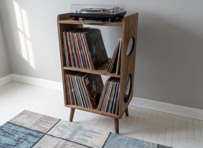 Op de afbeelding: Een houten platenspelerstandaard in mid-century modern stijl, met een draaitafel bovenop en vinylplaten opgeslagen op de planken. De standaard heeft een donkere houten afwerking en cirkelvormige uitsparingen.