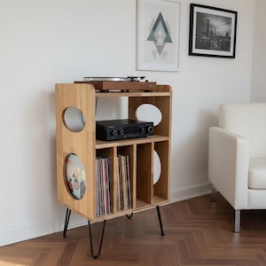 Puede incluir: Soporte de madera para tocadiscos con patas de metal negro. El soporte tiene varios estantes para discos y equipos de audio. Un tocadiscos y un amplificador están en la parte superior. El soporte está en una habitación con una pared blanca y un sillón blanco.