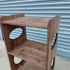 Op de afbeelding: Een houten plank met drie niveaus en een donkerbruine afwerking. De zijkanten hebben ovale uitsparingen en de bovenste plank heeft een verhoogde achterkant. De planken zijn gemaakt van massief hout, met de natuurlijke nerf.