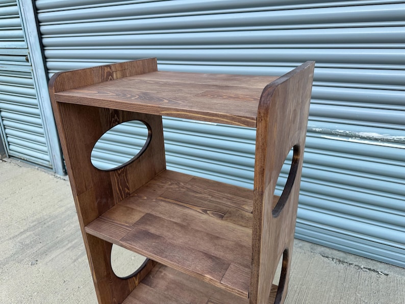 Op de afbeelding: Een houten plank met drie niveaus en een donkerbruine afwerking. De zijkanten hebben ovale uitsparingen en de bovenste plank heeft een verhoogde achterkant. De planken zijn gemaakt van massief hout, met de natuurlijke nerf.
