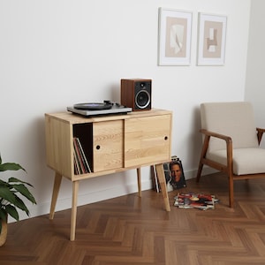 Puede incluir: Consola de tocadiscos de madera clara con puertas correderas, que contiene discos de vinilo. Un tocadiscos y un pequeño altavoz se encuentran encima. Un sillón beige y obras de arte enmarcadas están en el fondo.