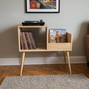 Puede incluir: Un mueble de madera clara para tocadiscos con un tocadiscos negro. El mueble tiene dos compartimentos, uno para discos de vinilo y otro para guardar álbumes. El mueble está sostenido por cuatro patas anguladas. Una imagen enmarcada cuelga de la pared encima.