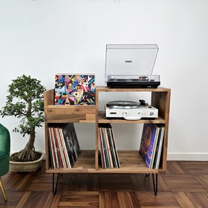 以下が含まれることがあります： 2台のターンテーブルとレコード収納を備えた木製レコードプレーヤースタンド。スタンドは天然木仕上げで、黒い金属製の脚が付いています。盆栽と緑のベルベットの椅子が背景にあります。
