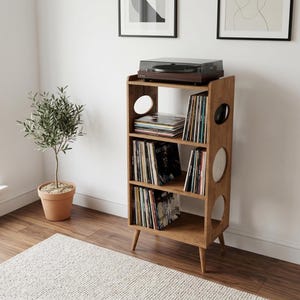 Puede incluir: Un mueble de madera para tocadiscos con tres estantes y recortes circulares. El estante superior contiene un tocadiscos. Los estantes están llenos de discos de vinilo. El mueble es de madera marrón claro y tiene patas anguladas.