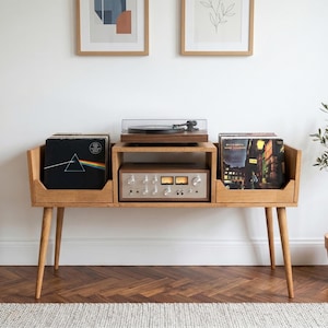 Puede incluir: Consola de discos de madera de estilo moderno de mediados de siglo con tocadiscos, amplificador y almacenamiento de discos. La consola tiene patas anguladas y un acabado de madera natural. Hay impresiones artísticas enmarcadas en la pared blanca.