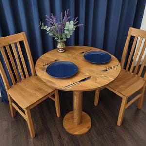 May include: A round wooden dining table with two matching chairs. The table is set with two blue plates, silverware, and a vase of flowers. The chairs have a slatted back design.