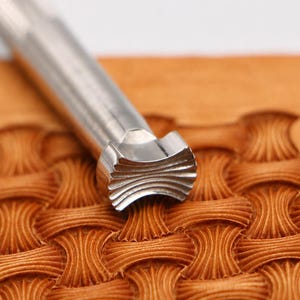 May include: A silver-coloured metal leather stamping tool with a cylindrical handle and a flared, grooved head. The tool is positioned on a piece of tan leather that features a repeating, embossed basketweave pattern created by the stamp.