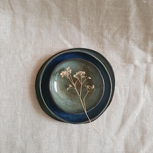Può includere: Vista dall'alto di tre piatti in ceramica in diverse tonalità di blu e verde, incastrati l'uno nell'altro. Un rametto di fiori secchi è appoggiato sui piatti, su uno sfondo di lino neutro. I piatti hanno un aspetto rustico e fatto a mano.