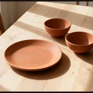 May include: A set of three terracotta-colored ceramic dishes consisting of one large flat circular plate and two smaller deep rounded bowls. The items feature a matte finish and a consistent earthy reddish-brown tone throughout the set.