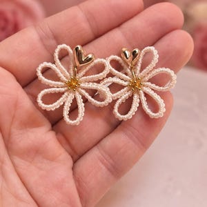 Puede incluir: Un par de pendientes de margaritas hechos a mano. Cada pendiente presenta un diseño de flor calada blanca con un acento de corazón dorado en el centro. Los pendientes se sostienen en la mano de una persona sobre un fondo rosa suave.