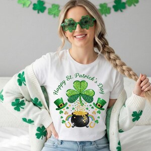 Może przedstawiać: Biała koszulka z motywem na Dzień Świętego Patryka. Projekt zawiera zieloną koniczynę, garnek złota i napis "Happy St. Patrick's Day". Projekt zawiera również zielone kapelusze i tęczę. Osoba nosi zielone okulary w kształcie koniczyny.