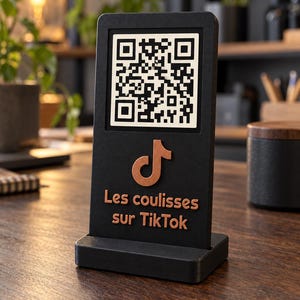Peut inclure: Un panneau vertical rectangulaire noir avec un code QR blanc en haut. Sous le code QR se trouve un logo TikTok couleur cuivre et le texte "Les coulisses sur TikTok" en lettres cuivrées. Le panneau est fixé sur une base rectangulaire noire.