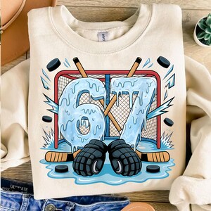 May include: Cream-colored sweatshirt with a hockey-themed graphic. The design features the number "67" in icy blue, a hockey net, pucks, hockey sticks, and hockey gloves. The graphic is a playful representation of hockey.