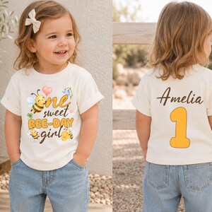 May include: A cream-colored short-sleeve t-shirt featuring a graphic of three bees, flowers, a heart, and the text "one sweet BEE-DAY girl" on the front. The back of the shirt displays the name "Amelia" in script and a large yellow number "1".
