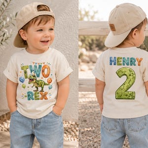 May include: A cream-colored short-sleeve t-shirt featuring a dinosaur graphic with the text "TWO-REX" on the front and the name "HENRY" above a large number "2" on the back, paired with a beige baseball cap and light blue denim jeans.