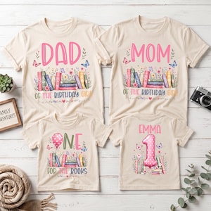 May include: Four beige t-shirts featuring colorful book and floral graphics. Two shirts display the text DAD and MOM OF THE BIRTHDAY GIRL. One shirt displays the text ONE FOR THE BOOKS with a balloon. One shirt displays EMMA and the number 1.