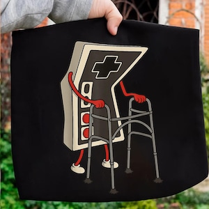 May include: Black tote bag featuring a cartoon illustration of a retro video game controller using a walker. The controller is beige and gray with red arms and legs, and the walker is gray.