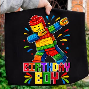 May include: Black fabric with a colorful graphic of a Lego-style figure dabbing. The figure is made of red, yellow, green, and blue blocks. The text "BIRTHDAY BOY" is below the figure in colorful block letters.