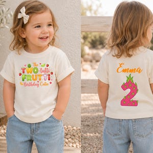 May include: A cream-colored short-sleeve t-shirt featuring a front graphic with the text The TWO-tutti FRUTTi Birthday Cutie surrounded by colorful fruit illustrations. The back displays the name Emma above a large pink dragon fruit number 2.