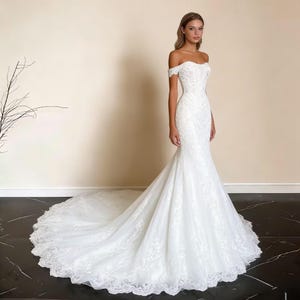 Puede incluir: Un vestido de novia blanco con hombros descubiertos y silueta de sirena. El vestido presenta intrincados detalles de encaje y una larga cola. La novia está de pie frente a un fondo neutro.