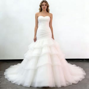 Puede incluir: Un vestido de novia blanco sin tirantes con escote corazón y falda con volantes en capas. El vestido presenta un corpiño ajustado y una falda fluida con volantes que se extiende en una cola. El vestido está hecho de una tela suave y fluida.