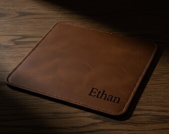 May include: A brown leather mousepad with the name "Ethan" printed in black. The square pad has stitched edges and a smooth surface. The mousepad is on a wooden surface.