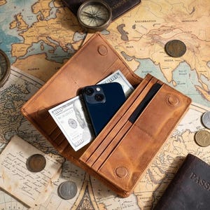 May include: A tan leather wallet, open to show a blue smartphone and US $100 bills. The wallet rests on a vintage map with a compass, coins, and a passport. The wallet has a snap closure.