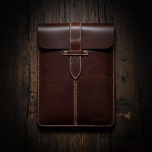Pode incluir: Uma capa de tablet de couro marrom com fecho de aba e uma alça vertical presa por um rebite de metal. A capa apresenta costuras brancas e o nome "LIAM" em relevo na parte inferior. O couro tem uma aparência rica e texturizada.