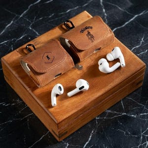 May include: Two brown leather AirPod cases with black carabiner clips and white AirPods on a wooden box. One case has a "Western" design and the other has a letter "K". The wooden box sits on a black marble surface.