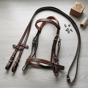 May include: A brown leather horse bridle with white stitching, metal hardware, and reins. The bridle is laid out on a light-colored surface, with small metal pieces, a wooden box, and a spool of thread nearby.