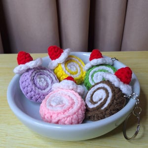 May include: A selection of crocheted Swiss roll keyrings in a white bowl. Each roll is a different colour, including purple, yellow, pink, green, and brown, with a white swirl and a red Santa hat on top. The keyrings are on a wooden surface.