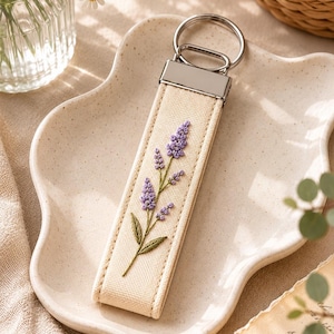May include: A beige fabric keychain with a silver metal ring and accents. The keychain features an embroidered lavender sprig in shades of purple and green. The keychain is displayed on a cream-colored, wavy-edged plate.