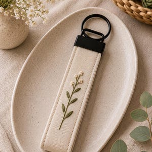 May include: Cream-colored keychain with a black metal ring and a rectangular fabric strap. The strap features an embroidered floral design in green and cream. The keychain is displayed on a beige oval plate.