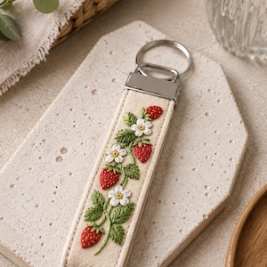 May include: Cream-colored keychain with a detailed embroidered design of strawberries, white flowers, and green leaves. The keychain has a silver-colored metal ring and a rectangular fabric strap. The design is centered on the strap.