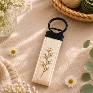 May include: Cream-colored keychain with a black metal ring and a floral embroidery design. The keychain features a rectangular strap with a stitched border and a delicate embroidered flower pattern in white and green.