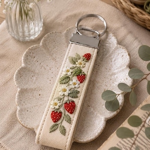 May include: Cream-colored keychain with a detailed embroidered design of red strawberries, green leaves, and white flowers. The keychain has a silver-colored metal ring. The design is centered on the cream-colored strap.