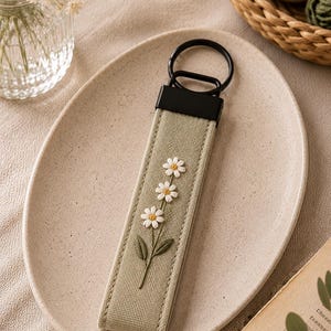 May include: A light green keychain with a black metal ring and a rectangular fabric strap. The strap features embroidered white daisies with yellow centers and green leaves. The keychain is on a beige plate.