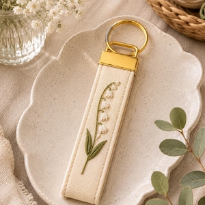 May include: A cream-colored keychain with a gold-tone metal clasp and ring. The keychain features an embroidered lily of the valley design in white and green. The keychain is displayed on a decorative plate.