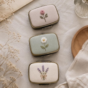 Könnte beinhalten: Drei rechteckige Metallkästchen mit Klappdeckeln, deren Oberseiten jeweils mit Stoff bezogen und handgestickten Blumenmotiven versehen sind. Das obere Kästchen zeigt eine rosa Rose, das mittlere ein weißes Gänseblümchen und das untere einen violetten Lavendelzweig.