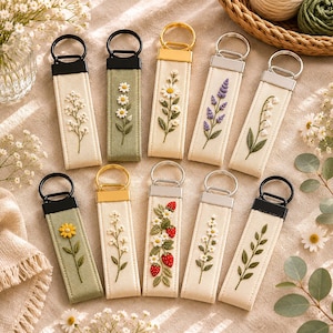 May include: Embroidered keyrings with floral patterns, such as daisies, lavender, and strawberries. Each keyring has a fabric strap with a metal ring and a contrasting black or gold top. The keyrings are presented on a neutral-toned surface.