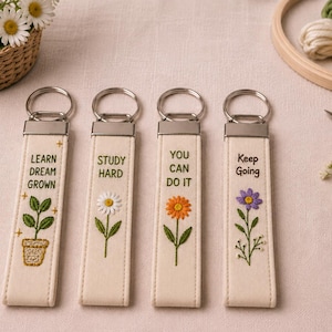 Könnte beinhalten: Vier rechteckige Schlüsselanhänger aus cremefarbenem Stoff mit silbernen Metallringen. Jeder zeigt gestickte Blumenmotive und Text: LEARN DREAM GROWN mit einer Topfpflanze, STUDY HARD mit einem weißen Gänseblümchen, YOU CAN DO IT mit einer orangefarbenen Blume und Keep Going mit einer violetten Blume.