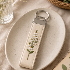 May include: Cream-colored keychain with a floral embroidery design. The keychain features a metal ring and a rectangular fabric strap. The strap is embroidered with a white and yellow flower design with green leaves. The keychain is resting on a cream-colored plate.
