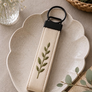 May include: A beige fabric keychain with a black metal ring and a black rectangular top. The keychain features an embroidered olive green leaf design. The keychain is on a beige scalloped plate.