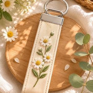 May include: Cream-colored keychain strap with embroidered white daisies and green leaves. The strap has a silver-tone metal top and a key ring. The keychain is on a wooden surface with daisy flowers in the background.