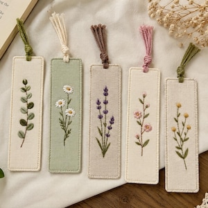Puede incluir: Cinco marcadores de tela bordados con diseños florales. Cada marcador presenta una flor diferente: eucalipto, margaritas, lavanda, flores rosas y flores amarillas. Cada marcador tiene una borla.