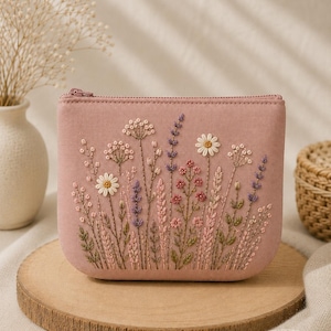 Könnte beinhalten: Ein rosafarbenes Täschchen mit Reißverschluss und gestickten Wildblumen. Das florale Design umfasst Gänseblümchen, Lavendel und andere zarte Blüten in Weiß-, Lila- und Rosatönen. Das Täschchen ist rechteckig und liegt auf einer Holzscheibe.