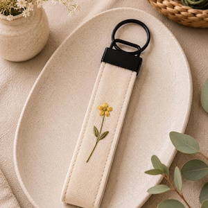 May include: Cream-colored keychain strap with a yellow embroidered flower and green stem. The strap has a black metal ring and a black rectangular piece at the top. The keychain is on a beige oval plate.