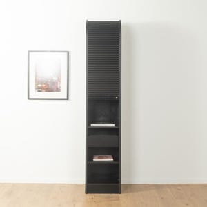 Pode incluir: Um armário alto e preto com uma porta de enrolar com ripas e várias prateleiras. O armário tem formato retangular e fica contra uma parede branca. Uma obra de arte emoldurada está pendurada na parede à esquerda.