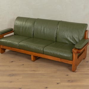 Könnte beinhalten: Ein Dreisitzer-Sofa mit Holzrahmen und olivgrünen Lederkissen. Das Sofa hat ein modernes Design aus der Mitte des Jahrhunderts mit Armlehnen und einem stabilen Holzsockel. Das Sofa steht auf einem Parkettboden.